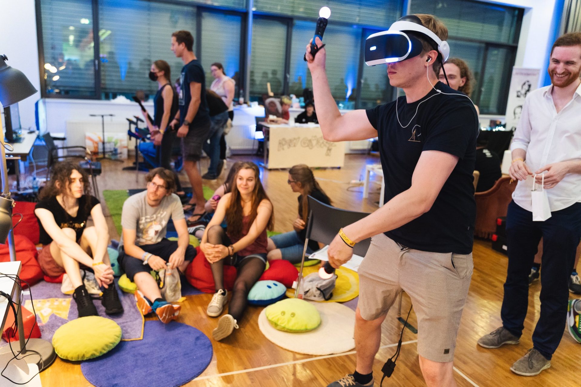 Ein Man beim Beat Saber spielen mit VR Headset und Motion Controllern, vor Publikum das auf Kissen sitzt.